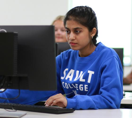 Student sitting at computer in class wearing blue Sault College sweater