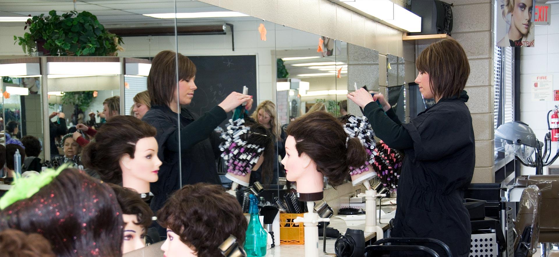 One female hairstylist student practices on a wig in front of a mirror. 