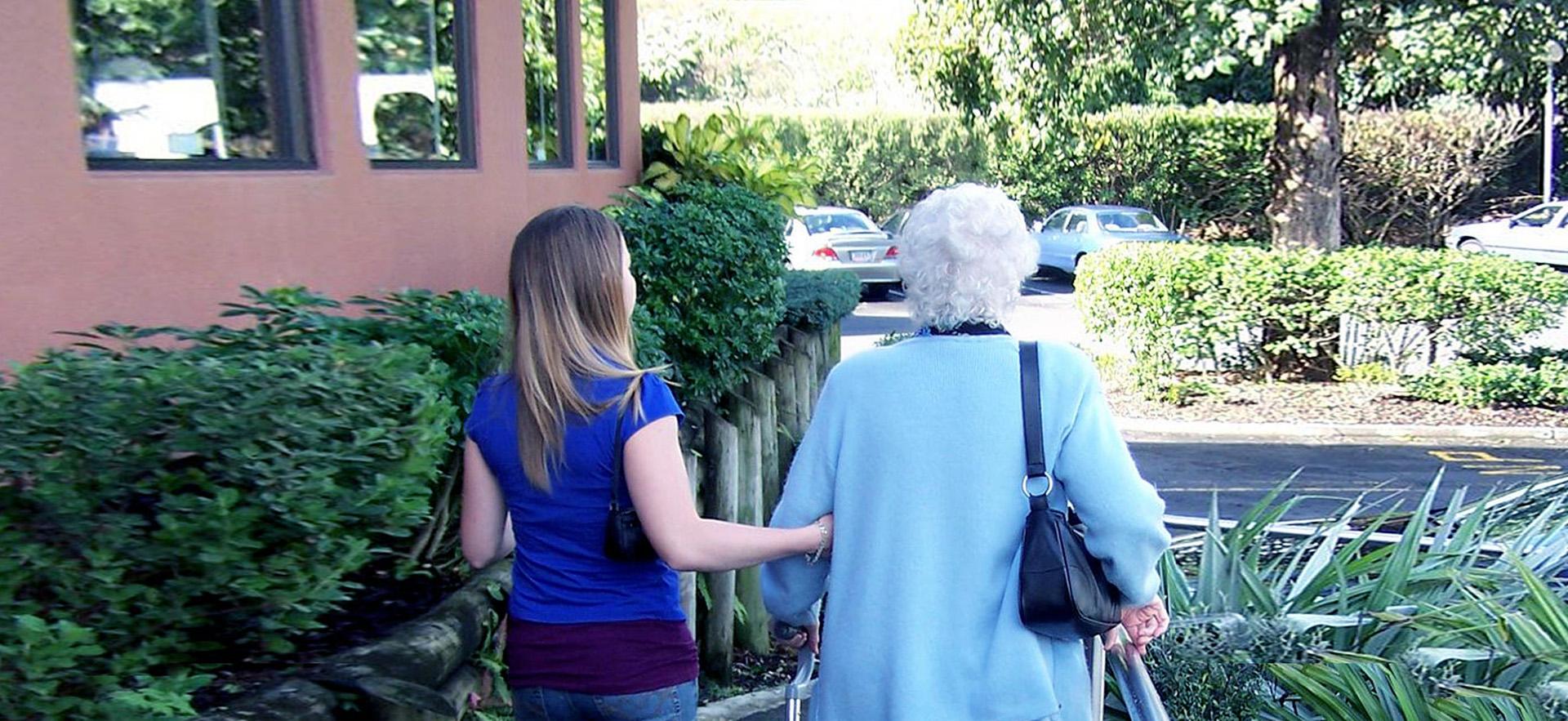 A young female helps an elderly woman walk down a sidewalk. 