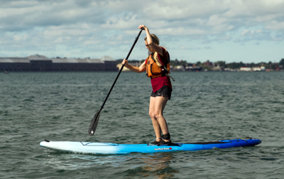 female wearing a life jacket on a standup paddleboard rowing in the water with bridge, trees and buildings in background