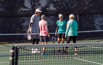 four people laughing while chatting and standing to the middle of the net on a tennis court