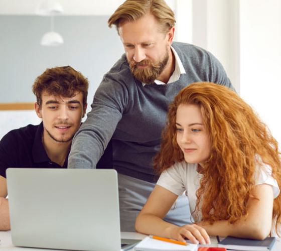 Adult standing between two students sitting at a desk and pointing to laptop screen