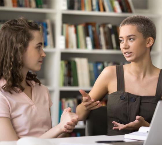 Two female students sitting together in library talking at a table