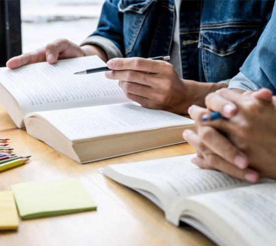 Two students showing just their hands sitting at a table with books and a pen