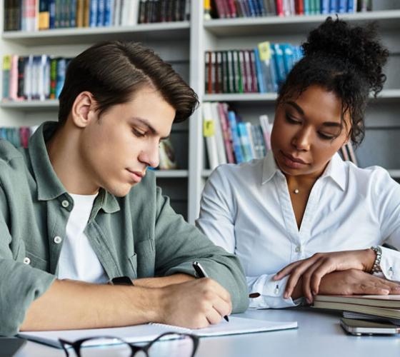 Two people sitting together looking at work on a table with bookshelf behind