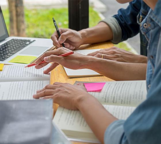 Two people seated at a desk together shown arms only with books and computer