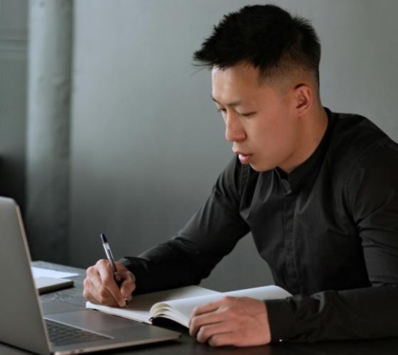 Man sitting at table at home with laptop and pen in hand writing in notebook
