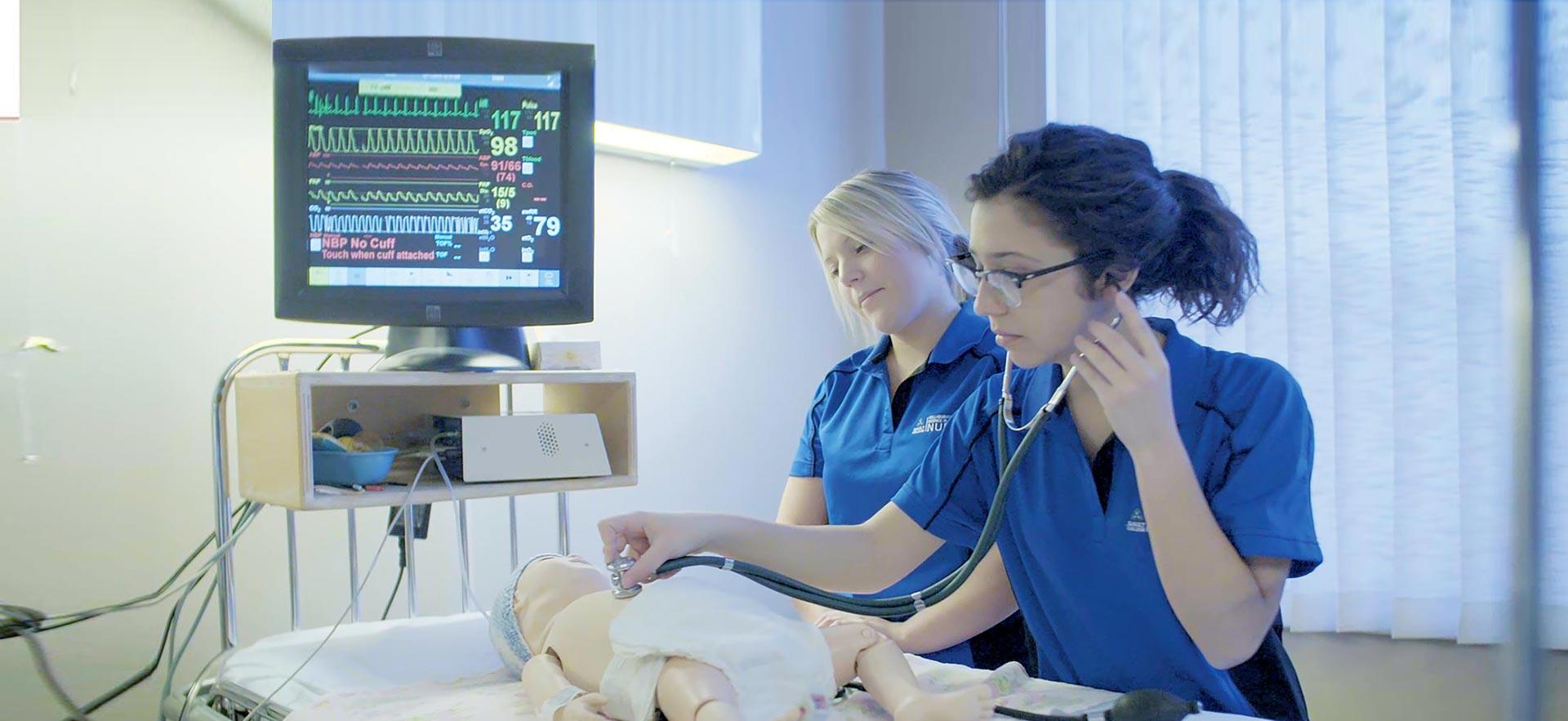 Two female Collaborative Bachelor of Science in Nursing students check the heart rate of a infant healthcare simulation mannequin. 