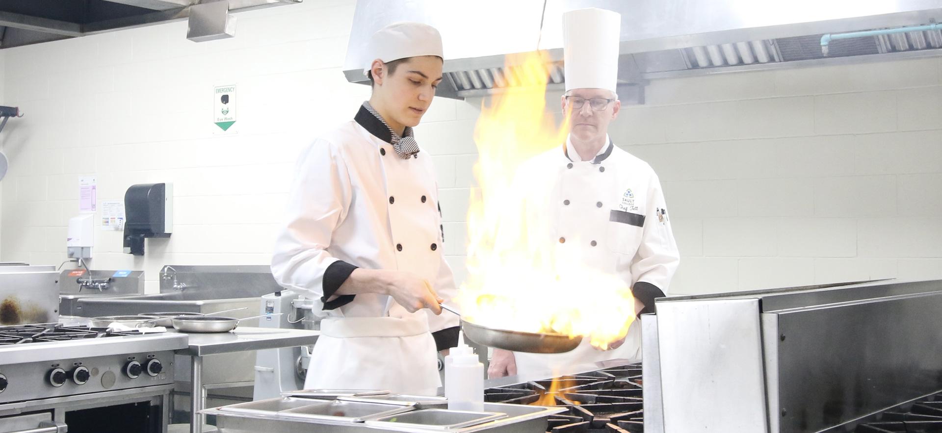 Two men in chef coats and hats in a professional kitchen with one with a pan and flambe in hand and the other overseeing